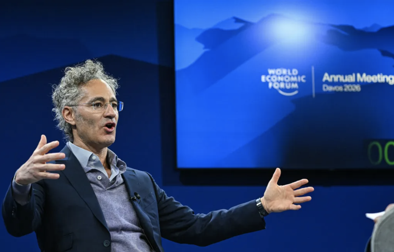 Palantir CEO Alex Karp speaking with both hands raised on stage at the World Economic Forum Annual Meeting in Davos, with the event backdrop behind him.